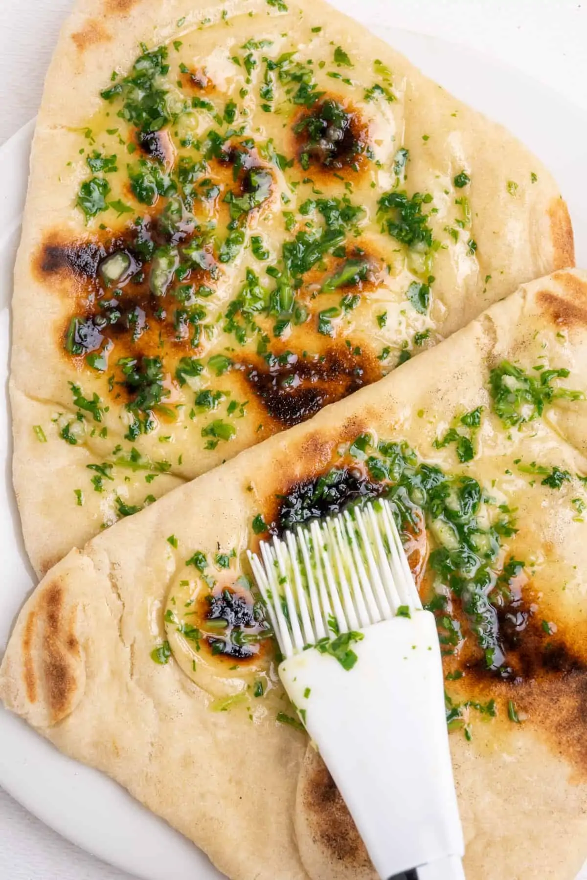 White flatbreads with dark brown, charred spots from cooking in a hot cast iron pan are brushed with melted butter infused with pressed garlic and chopped fresh coriander.