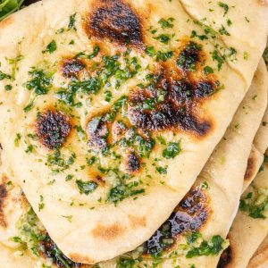 A stack of naan breads, with dark brown spots ono the top and brushed with garlic and coriander butter.