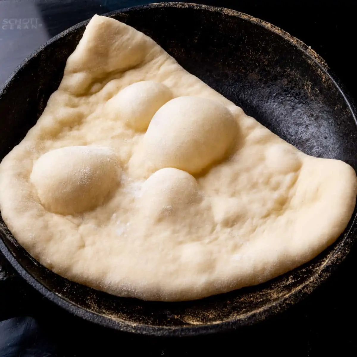 A flat piece of dough in a cast iron skillet. The top of the bread has puffed up into several large bubbles.