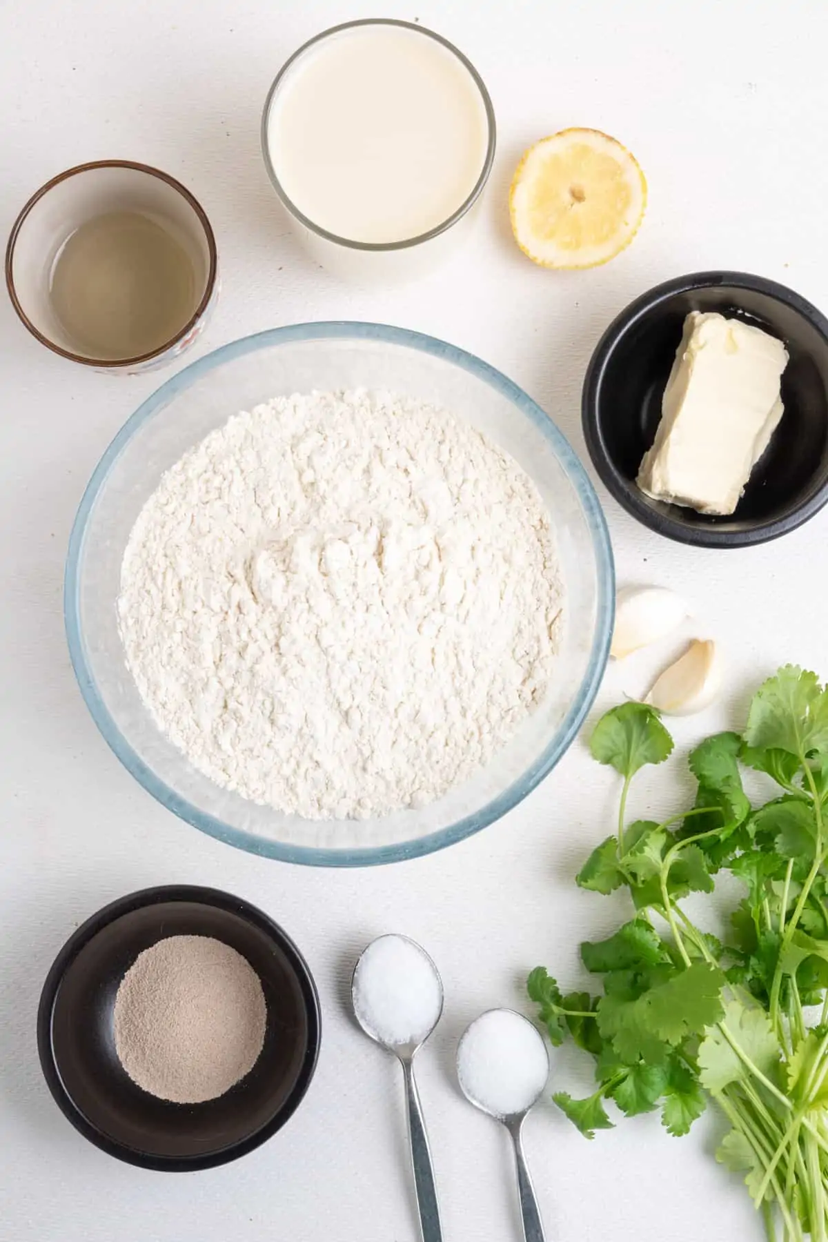 The ingredients for garlic and coriander naan: a bowl of plain flour surrounded by smaller pots of dried yeast, soy milk, vegetable oil, as well as a teaspoon each of sugar and salt, a slice of lemon, and some vegan butter, fresh coriander and two garlic cloves.