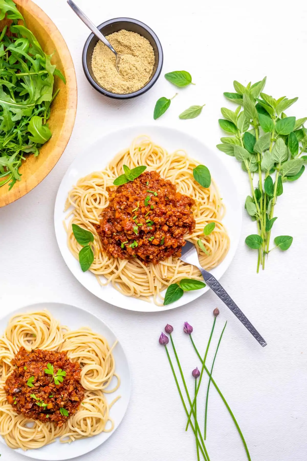 Top down view of a dining table. Two plates of spaghetti bolognese, a bunch of fresh herbs and chives, a small pot of vegan parmesan and a bowl of rocket.