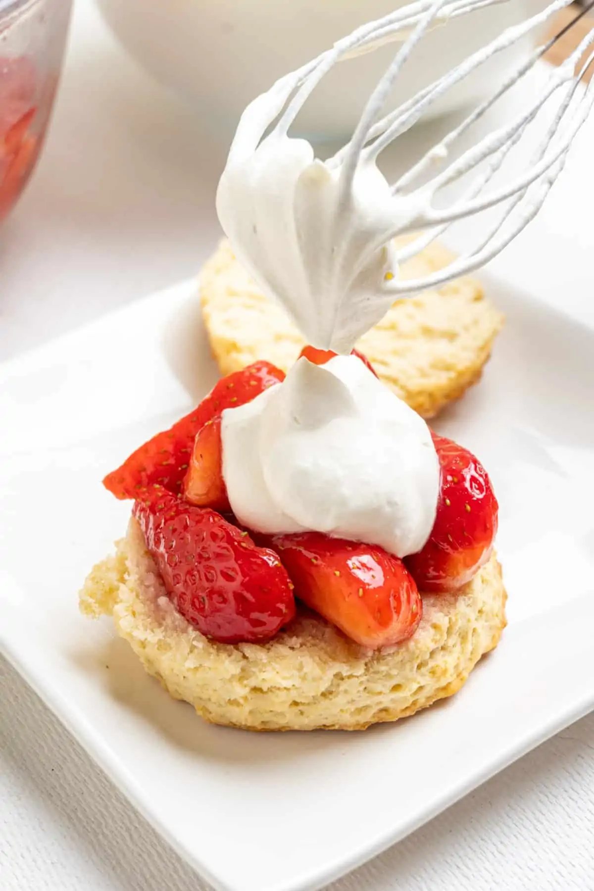 A whisk is dropping a dollop of whipped cream onto a bisuit topped with juicy red strawberries.