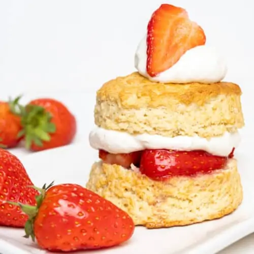 Two layers of golden baked biscuit are filled with juicy strawberries and whipped cream. The top biscuit is topped with more cream and a fresh strawberry.