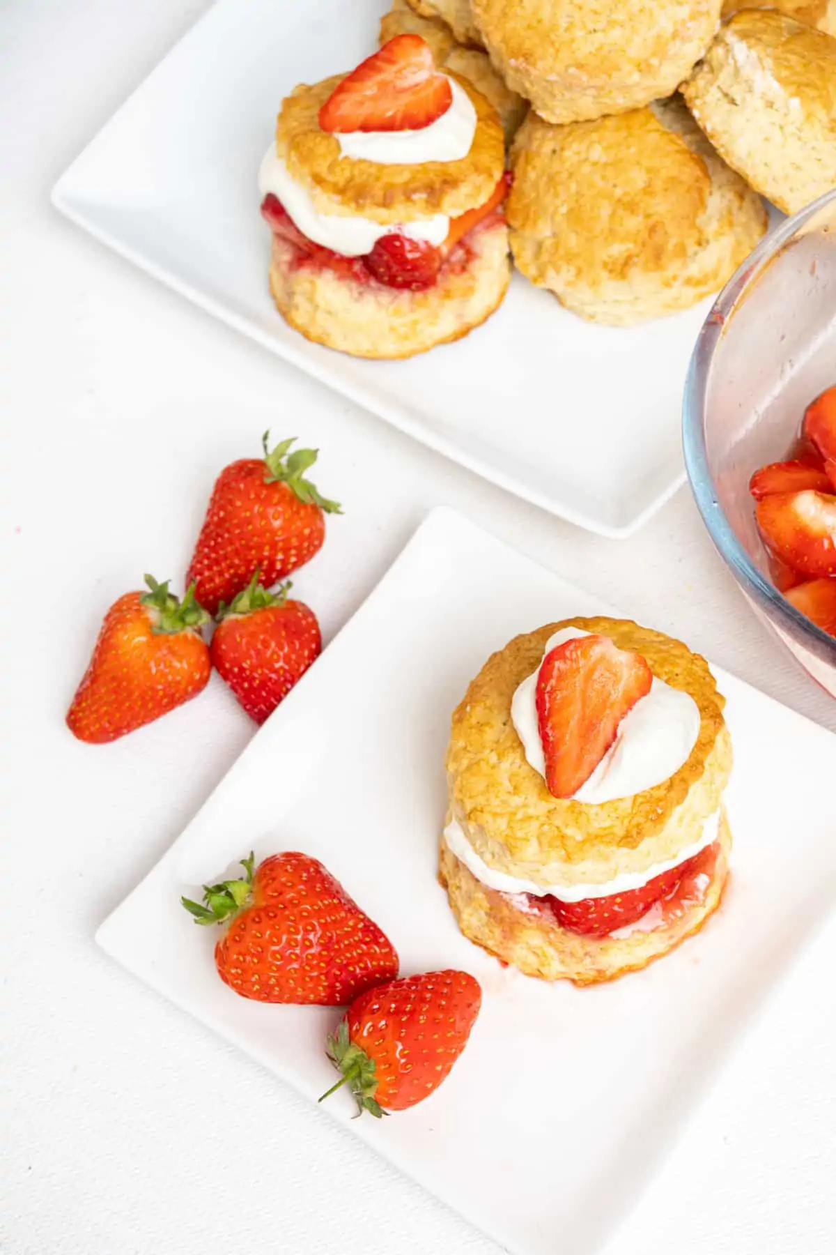 A tall biscuit with a cream and strawberry filling and topping served on a plate with fresh strawberries. More shortcakes on a serving platter in the back.