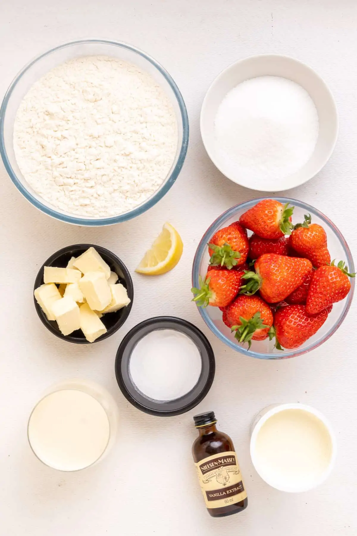 The ingredients for vegan strawberry shortcake: Strawberries, white flour, sugar, baking powder, cubes of vegan butter, soy milk, vegan whipping cream, vanilla extract and a small piece of lemon.