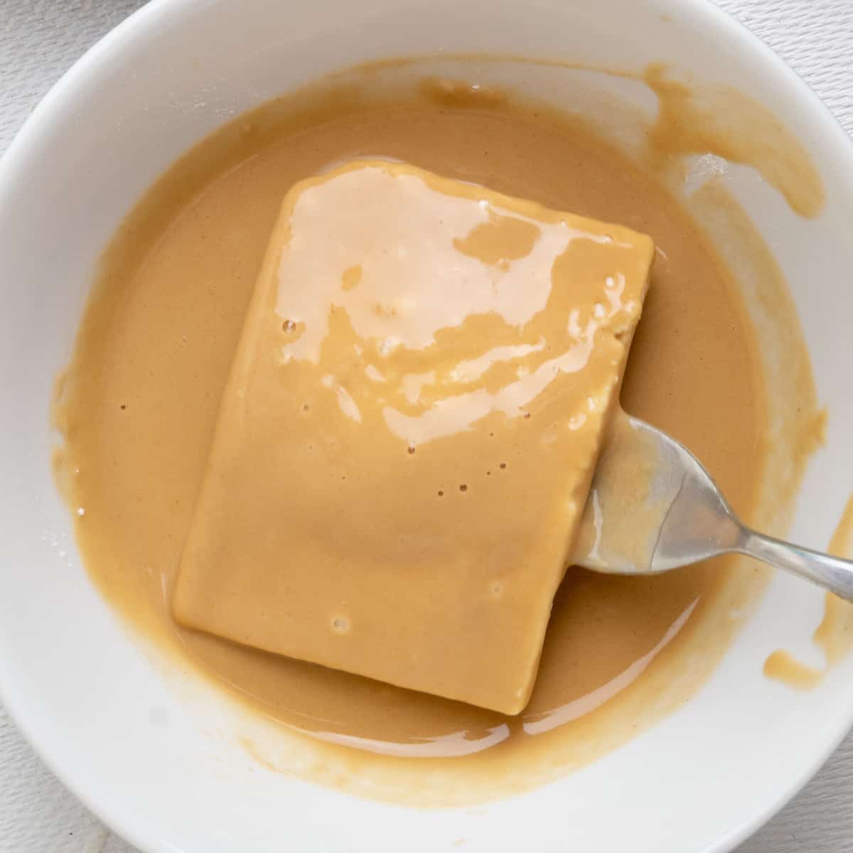A slice of tofu covered in a light orange brown batter lifted from a bowl on fork.