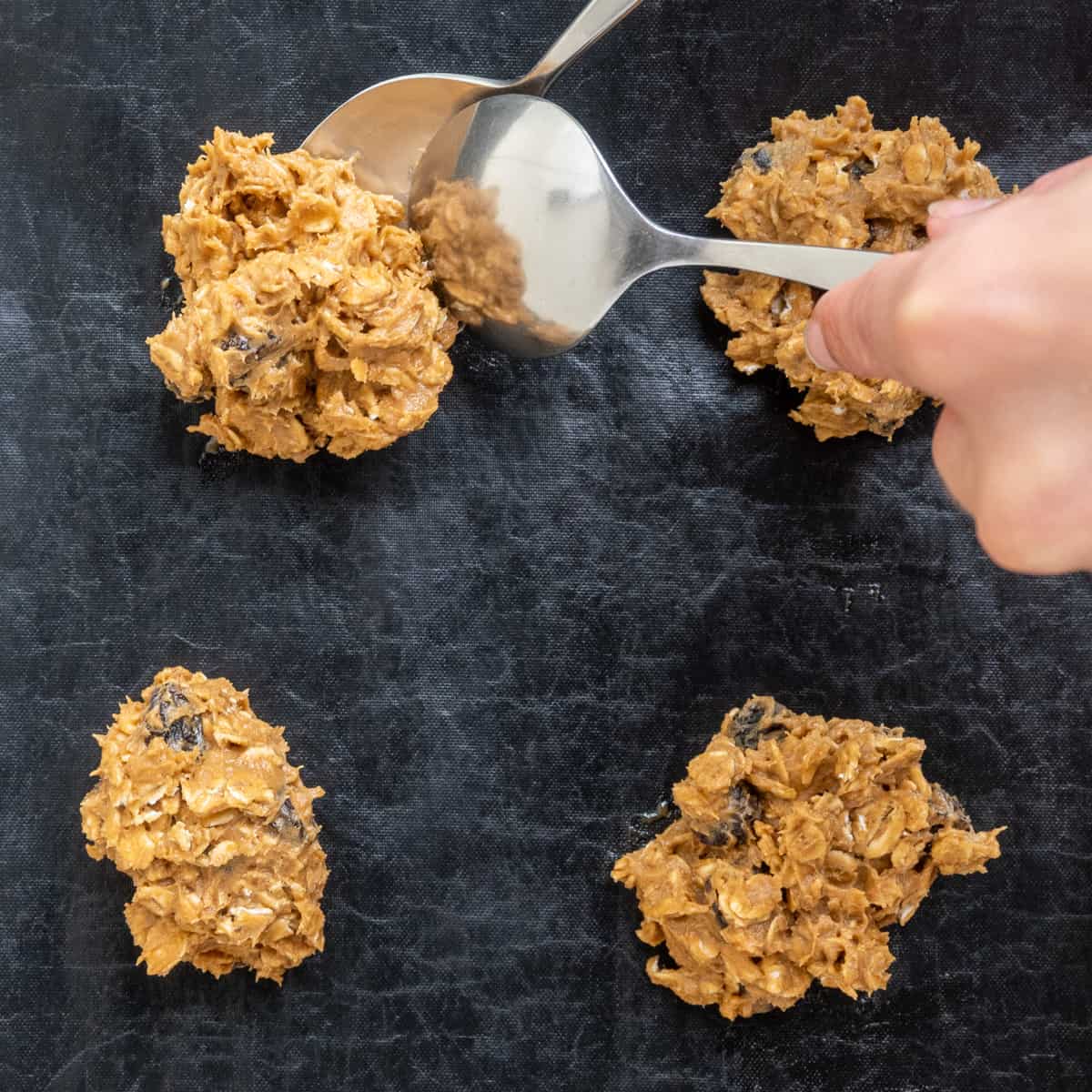 Scoops of the sticky cookie dough are placed on a lined baking tray using two tablespoons.