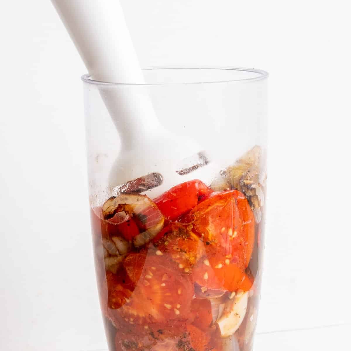 A handheld blender is turning the roasted vegetables into a sauce in a jug.