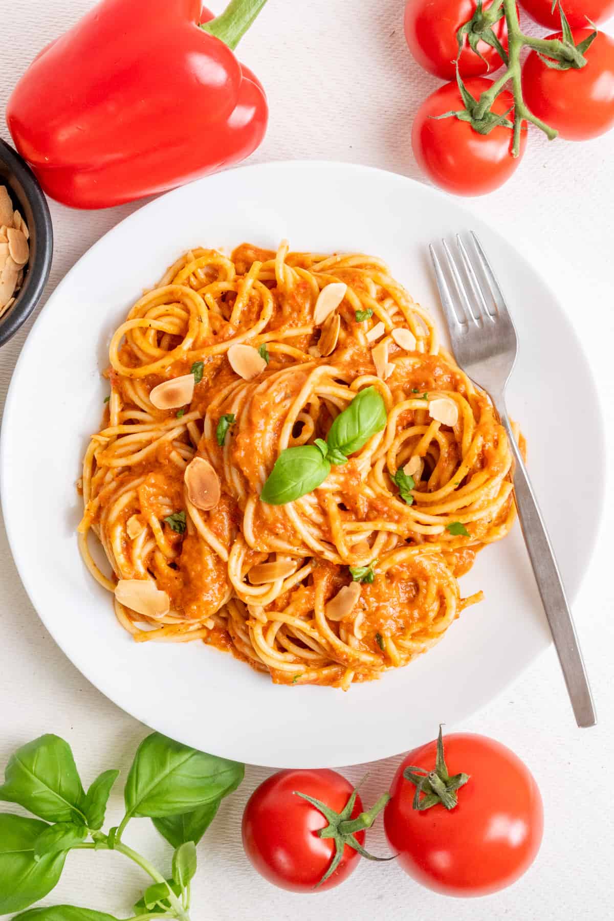 A plate of pasta with red pepper and tomato sauce, next to fresh tomatoes, a red pepper and herbs.