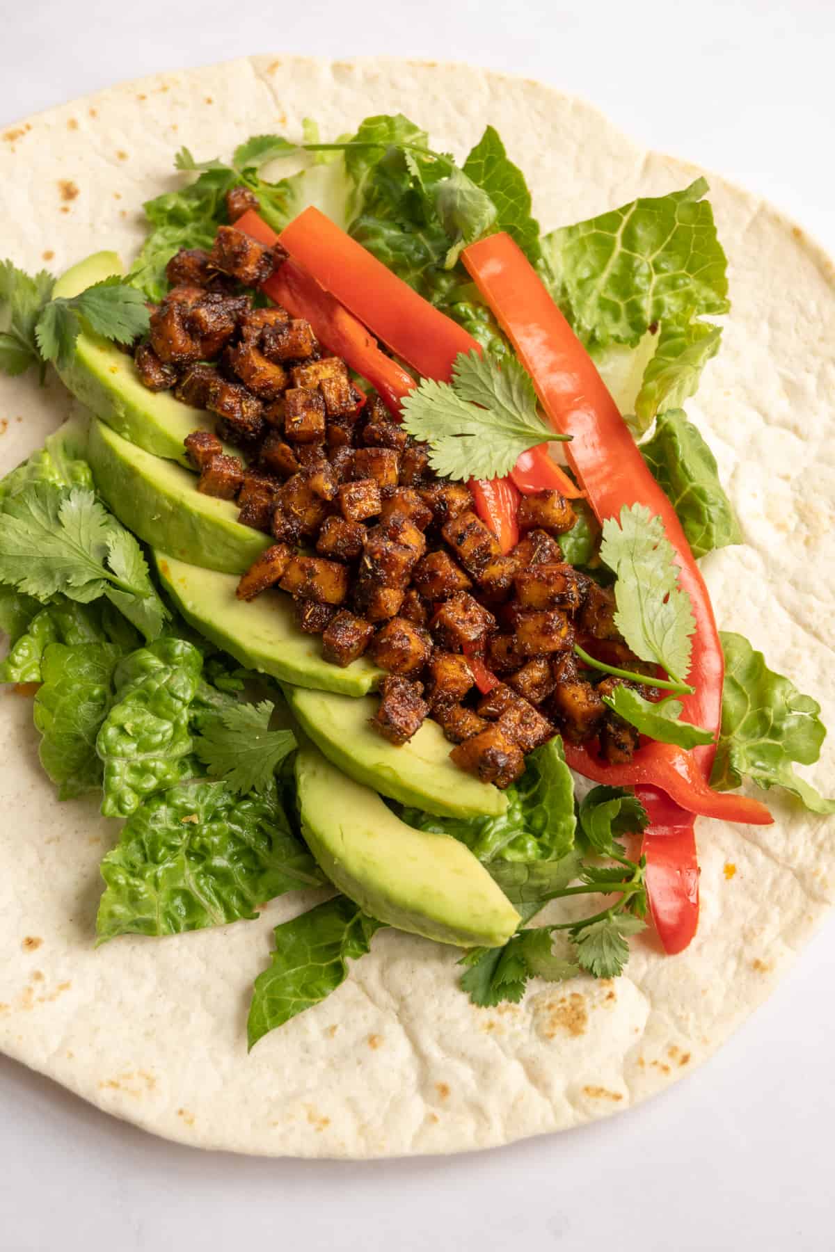The fillings for a tofu wrap are assembled on a tortilla: shredded lettuce, avocado, red pepper, spicy tofu cubes and fresh cilantro.