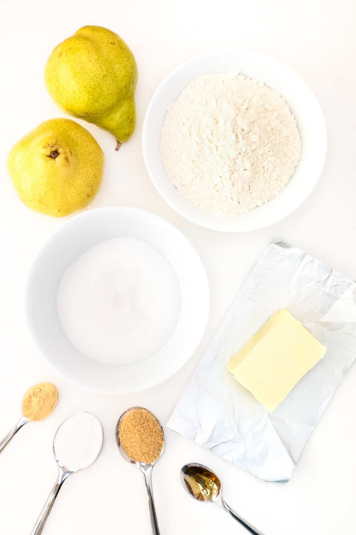 The ingredients for the vegan pear cake recipe laid out on a white surface. Pears, white flour, sugar, vegan butter, baking powder, ground ginger, vanilla extract and demerara sugar.