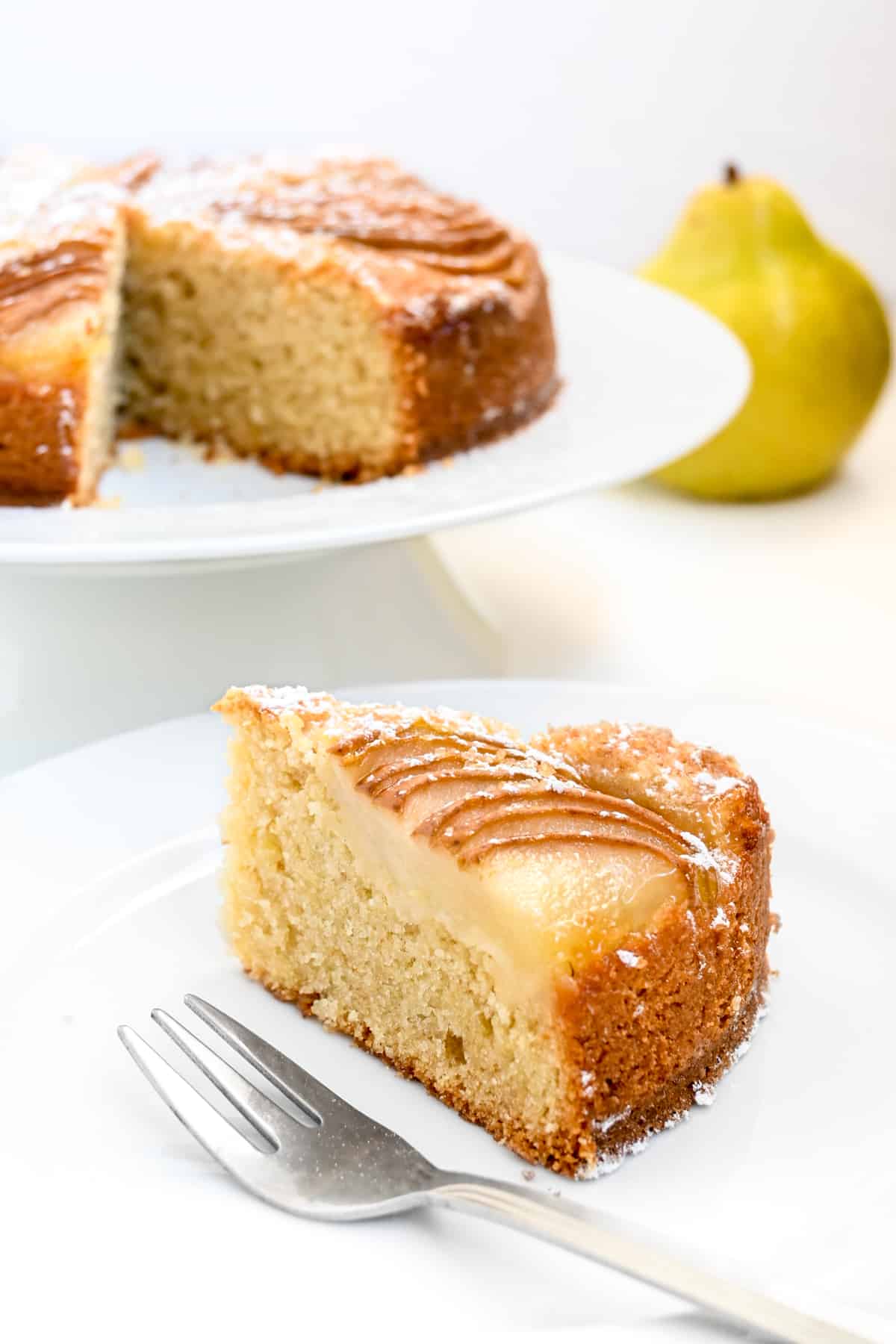 A slice of pear cake on a plate in front of the cake on the cake stand.