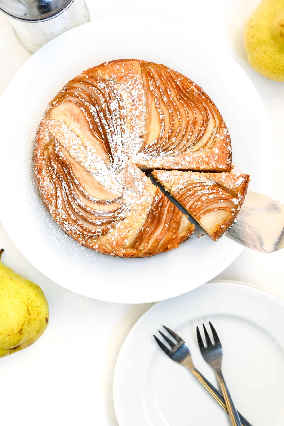 A slice of cake topped with pears and dusted with icing sugar is lifted and transferred onto a plate.