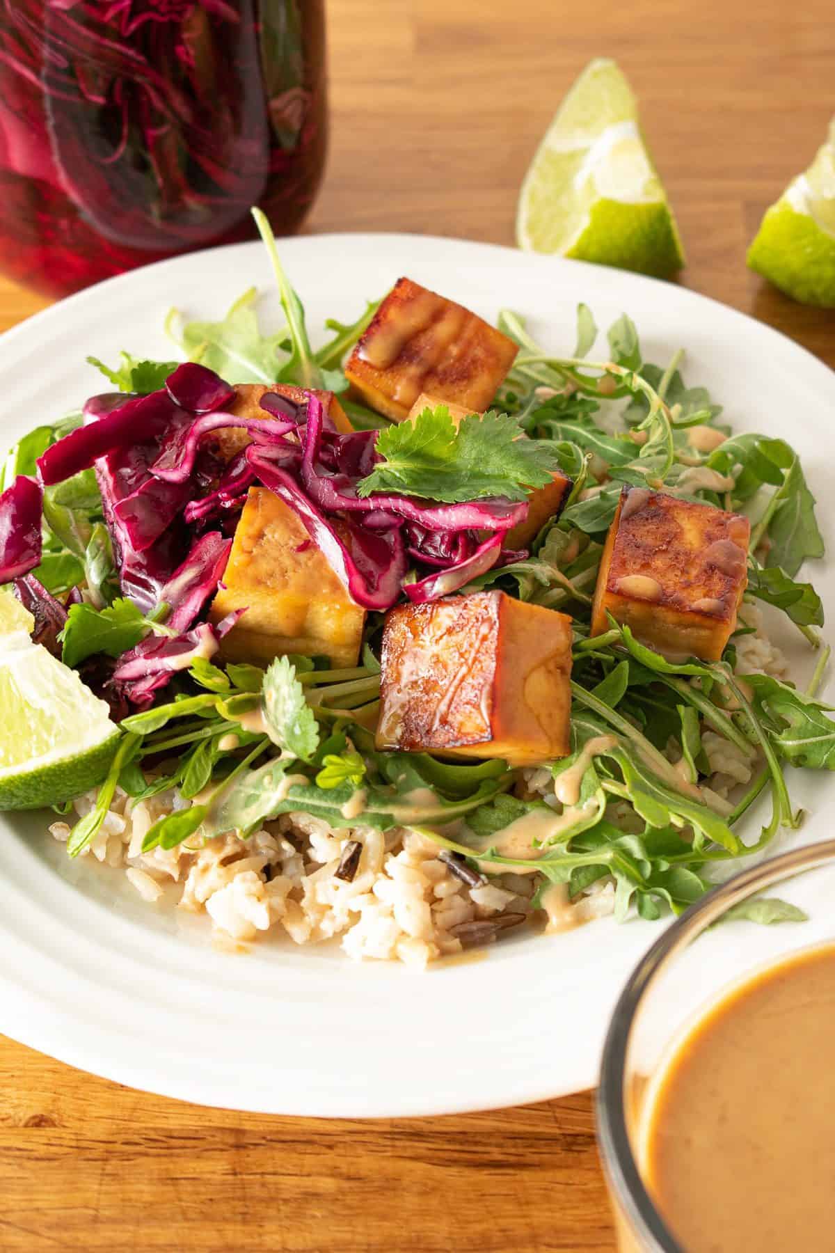A bowl of tofu with peanut sauce, greens, brown rice and pickled cabbage.