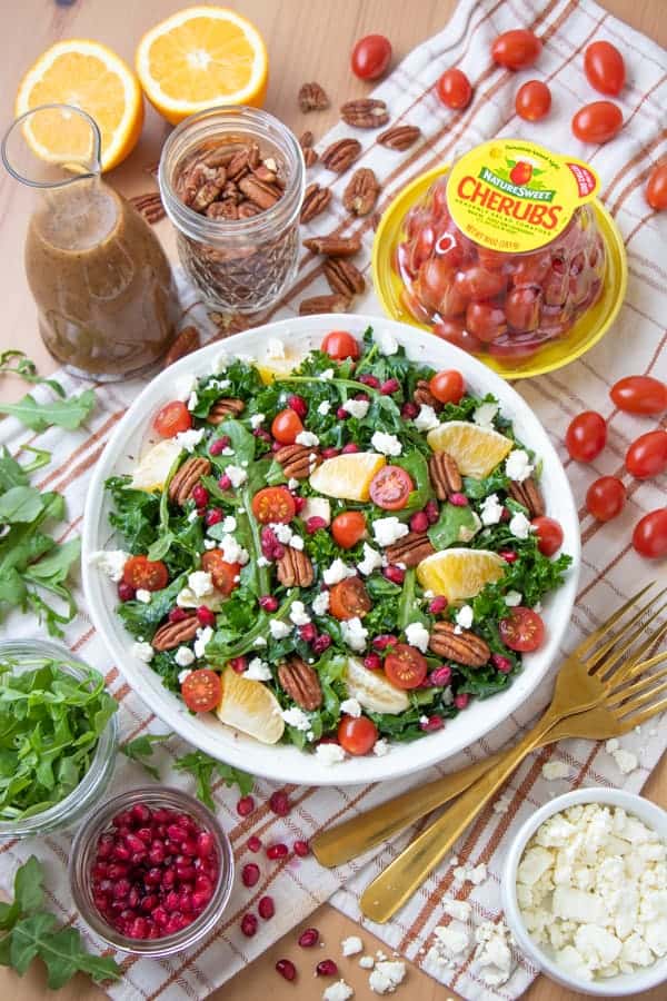 A bowl of kale salad topped with vegan feta, oranges, tomatoes, pomegranate and pecans.