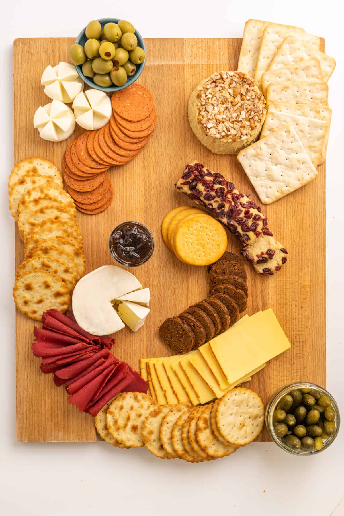 Arranged on a wooden board, three types of crackers, various vegan cheeses, ready-to-eat vegan deli slices and small bowls of olives, pickles and chutney.