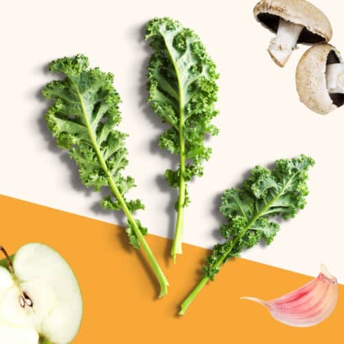 Kale leaves on an orange and cream backdrop, surrounded by other ingredients - apple, mushrooms and garlic.