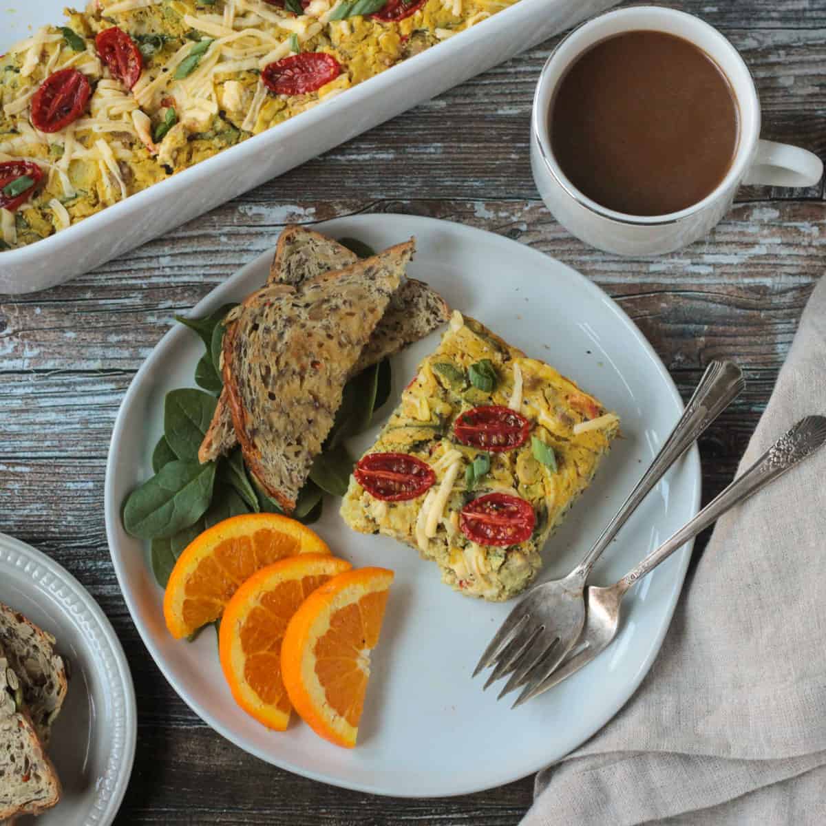 A slice of vegan breakfast casserole on a plate with toast and oranges.