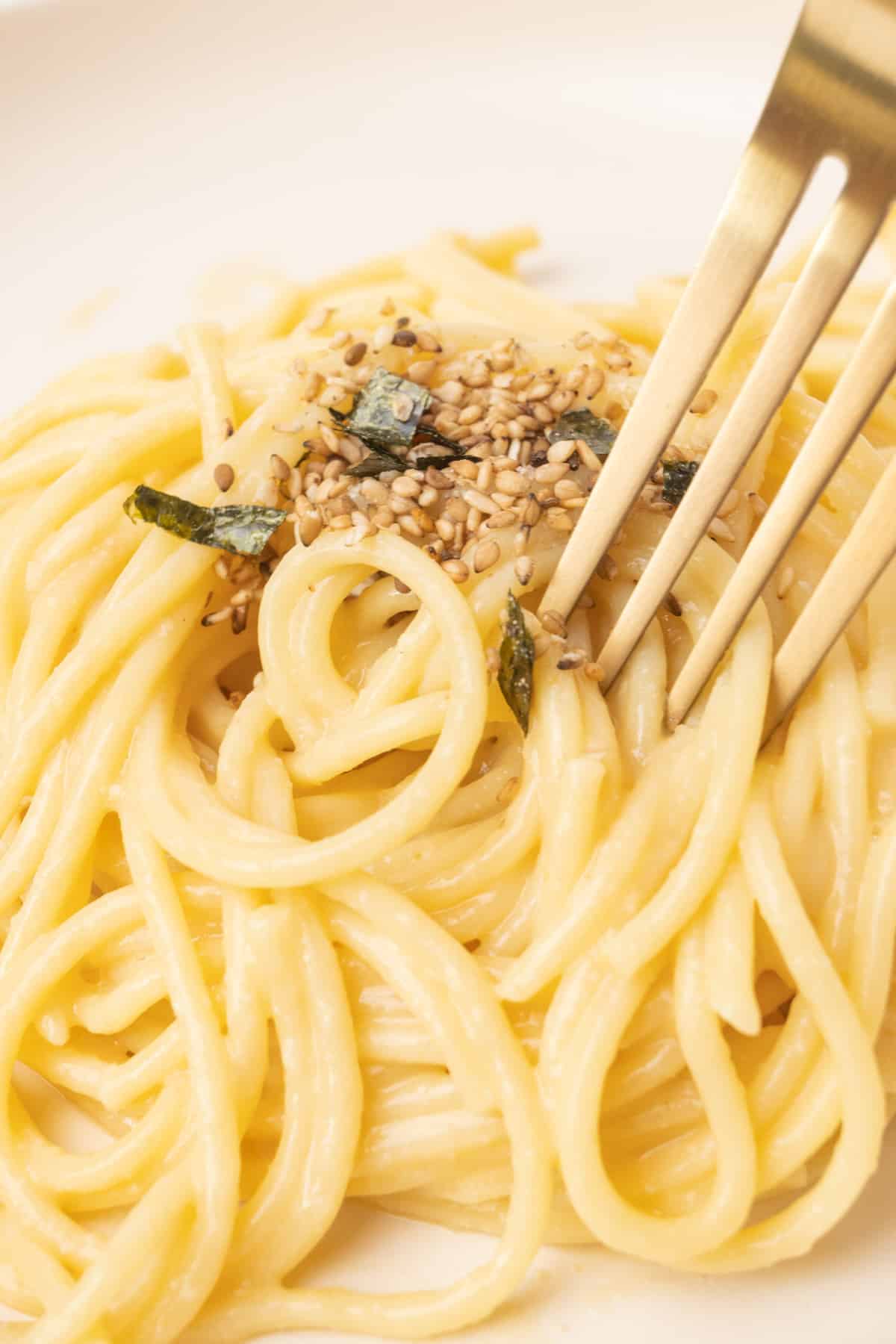 A fork digging into spaghetti coated in a creamy and glossy sauce and topped with sesame seeds and seaweed flakes.