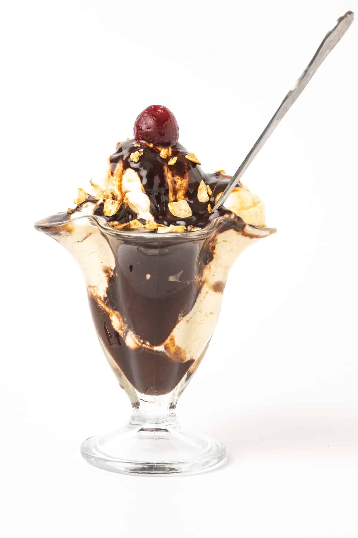 A vegan sundae with vanilla ice cream, hot fudge sauce, candied nuts and a cherry on top, served with a long dessert spoon..