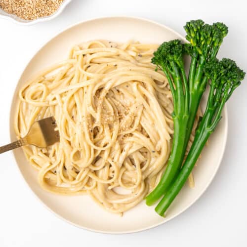 A fork digging into a plate of pasta in a glossy, creamy sauce served with tenderstem broccoli and a sprinkle of toasted sesame.