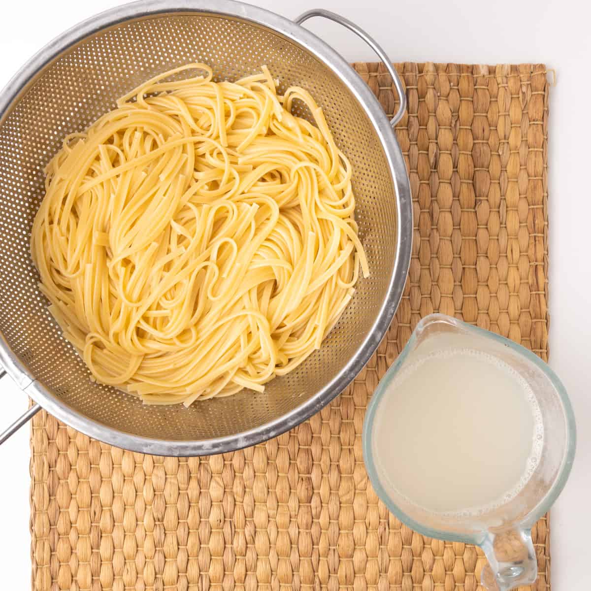 Cooked linguine pasta in a metal colander and a jug of starchy pasta cooking water.