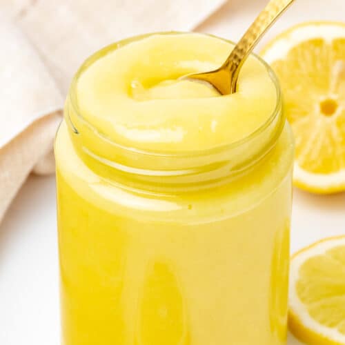 A golden teaspoon dipping into a jar of smooth, thick and creamy lemon curd.