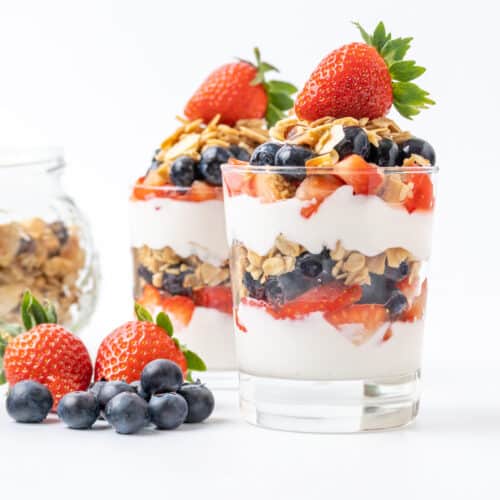 Two parfaits in glasses, made up of layered sweetened vegan yogurt, fresh strawberries and blueberries and vegan granola, topped with a whole strawberry.