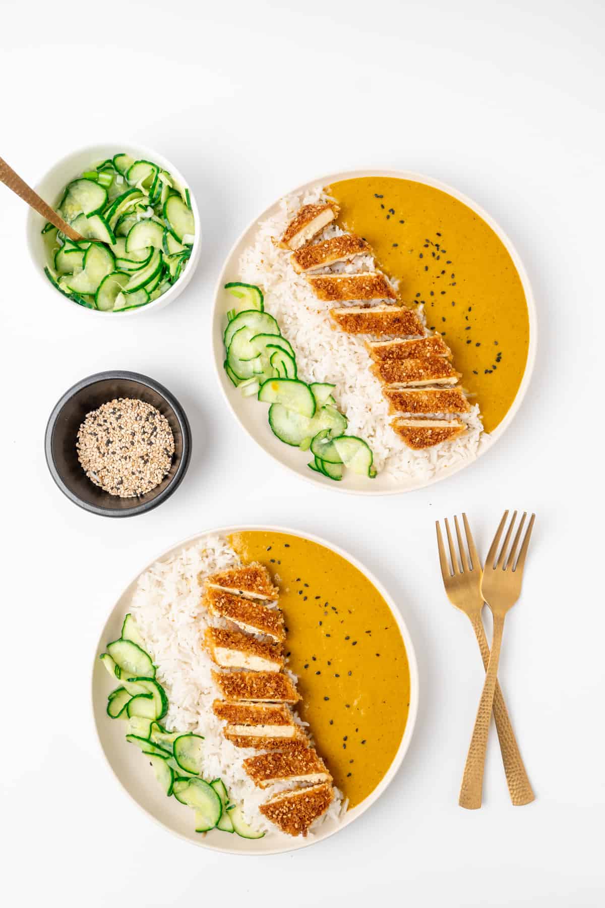 Two plates of vegan katsu curry, with cucumber pickles and sesame seeds on the side.