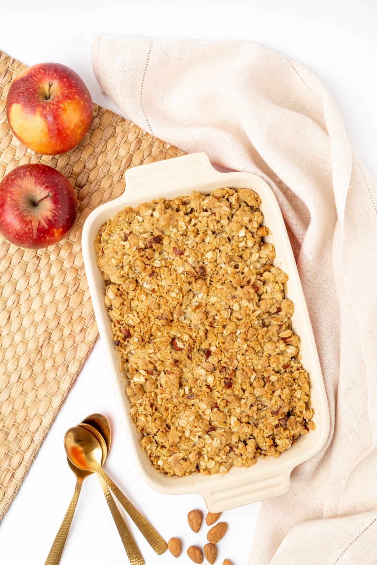 Apple crisp in a ceramic baking dish, next to spoons, a linen and almonds.