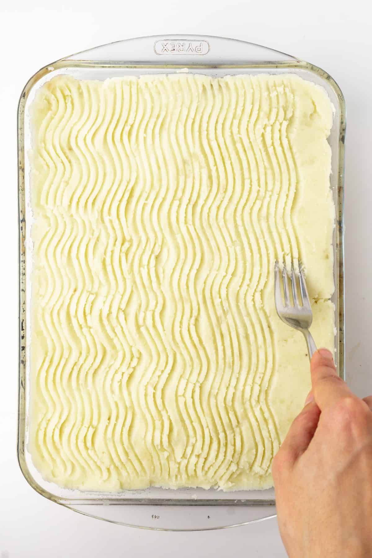 a fork carving waved lines into mashed potato in a rectangular baking dish.