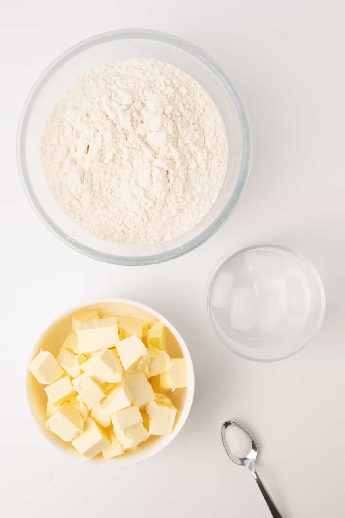 A bowl of flour, some cubed vegan butter, a glass of water with ice, and a small spoon of salt.