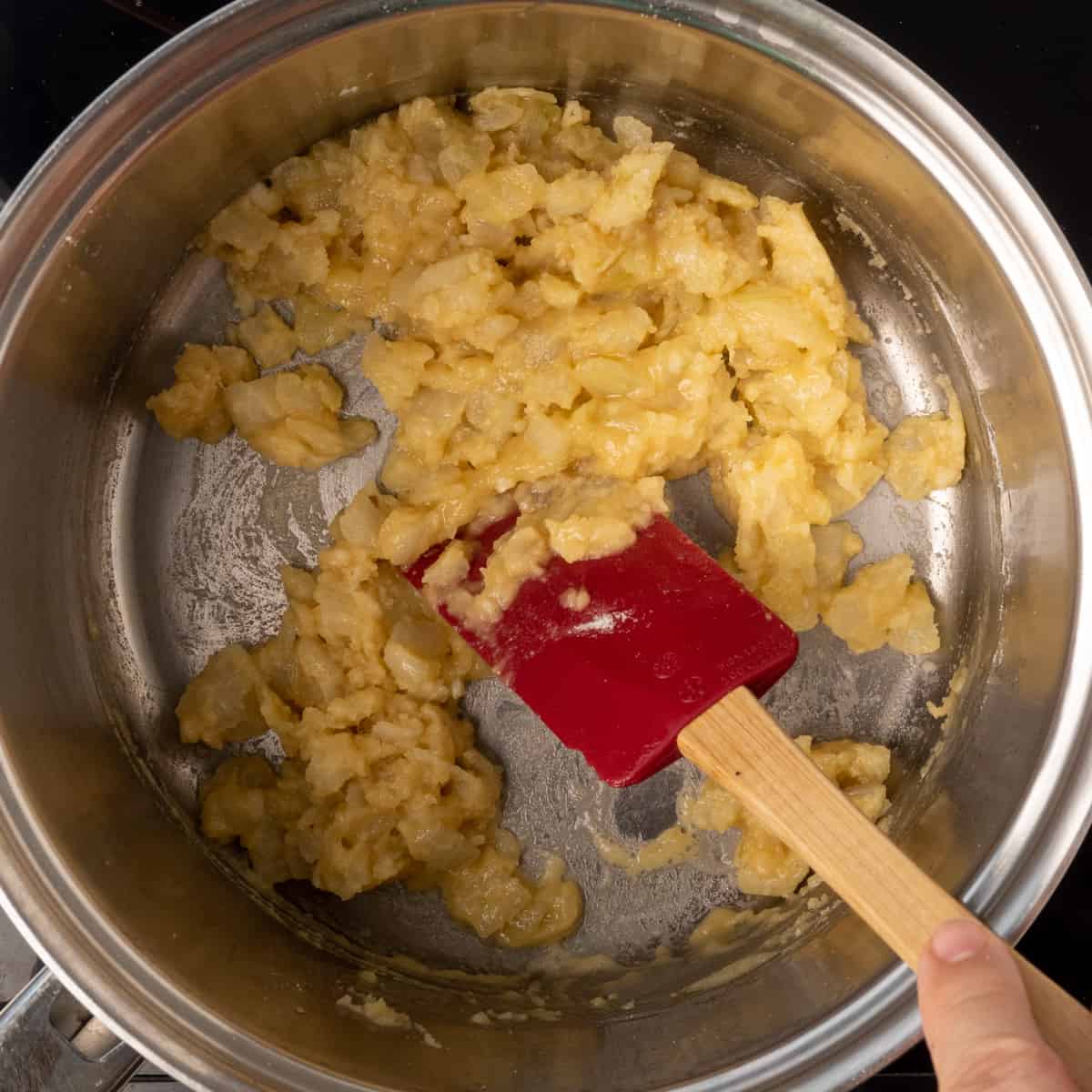 A silicone spatula stirring a thick, floury paste with diced onion in a saucepan.