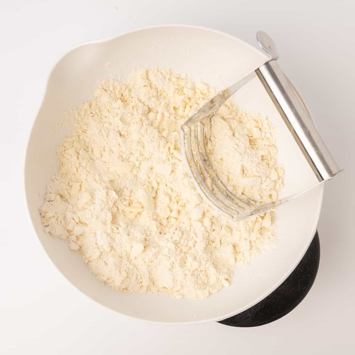 A crumbly flour and fat mixture in a bowl with a pastry cutter.