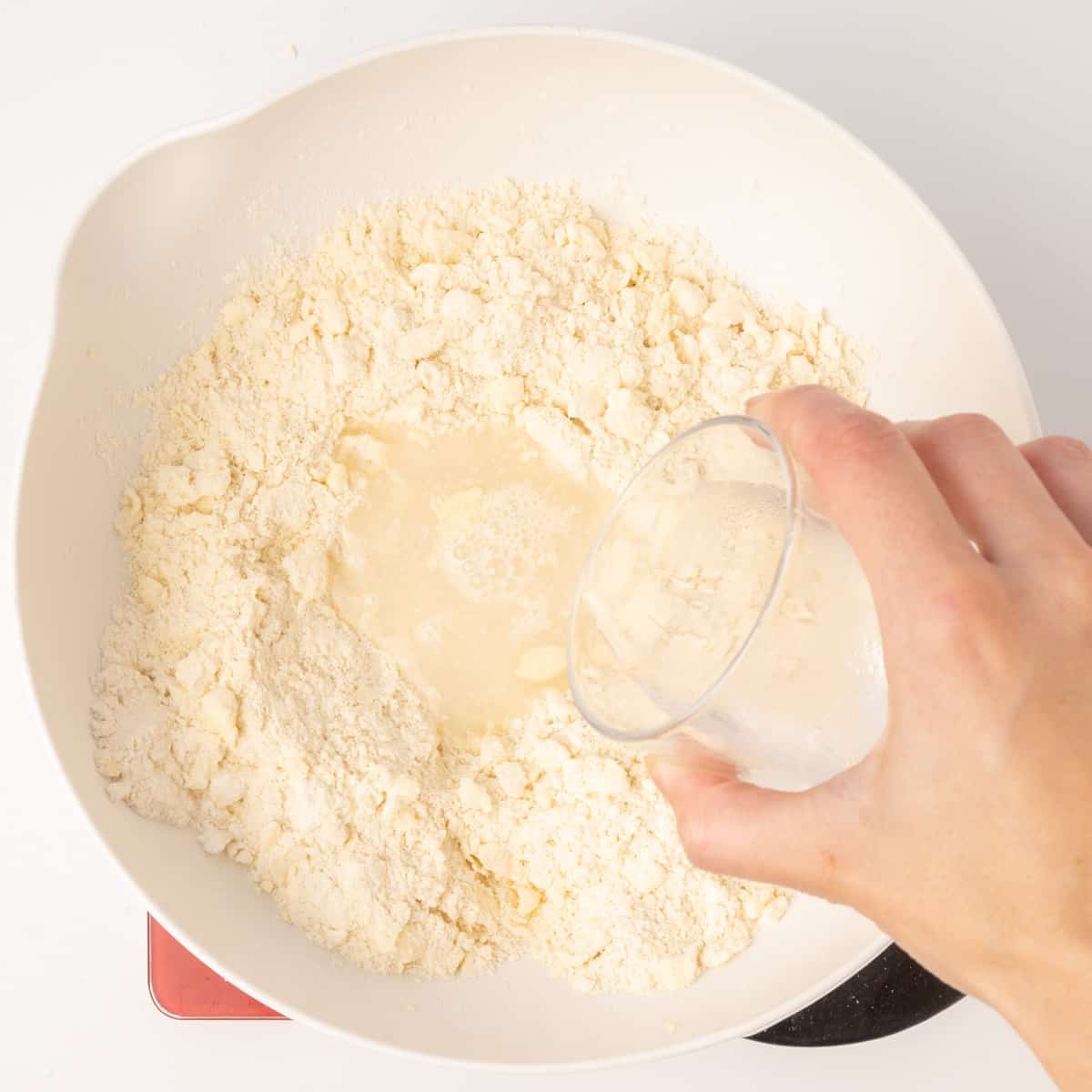 Cold water is poured from a glass into a bowl with a crumbly flour mix.