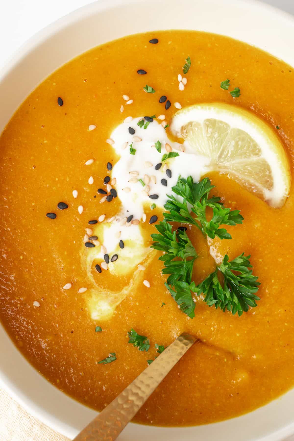 A bowl of blended carrot and red lentil soup topped with a spoonful of vegan creme fraiche, chopped parsley, sesame seeds and a slice of lemon.