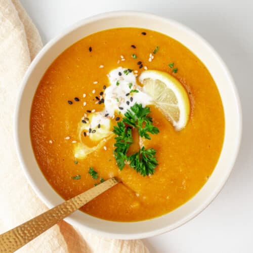 A bowl of orange coloured soup garnished with vegan creme fraiche, fresh herbs, sesame seeds and a slice of lemon.