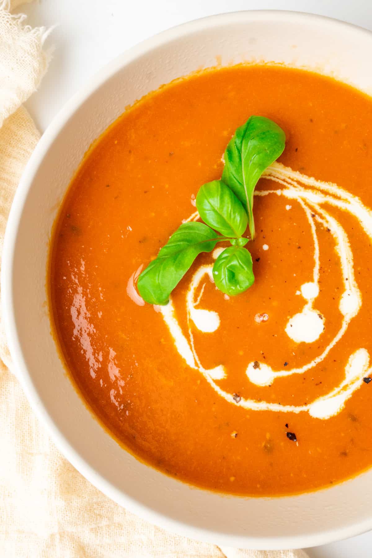 A bowl of tomato soup garnished with a drizzle of vegan cream and basil leaves.