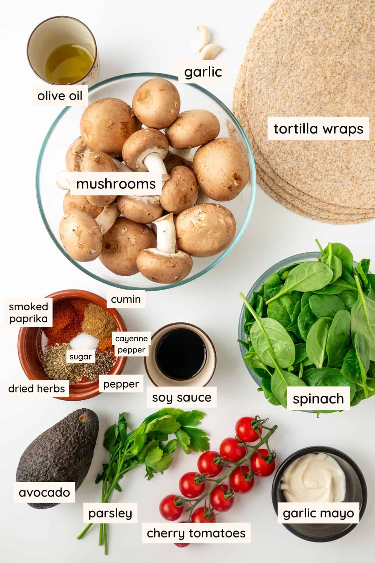 Ingredients o n a white surface: mushrooms, olive oil, garlic, tortilla wraps. smoked paprika, cumin, cayenne pepper, pepper, dried herbs, soy sauce, spinach, avocado, parsley, cherry tomataoes and garlic mayo.