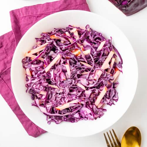 Red cabbage and apple slaw in a white salad bowl, the colour of the slaw matching the purple napkin underneath.